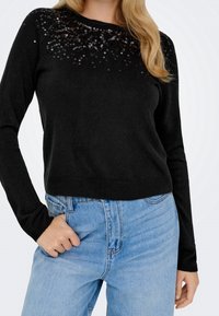 Femme portant un pull noir à manches longues avec des sequins près du col et un jean taille haute bleu clair, main dans la poche.