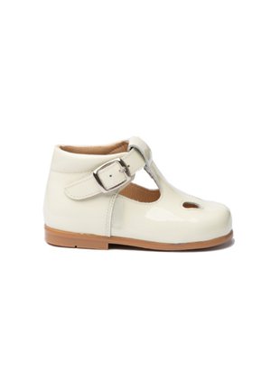 Zapato tipo Mary Jane de charol blanco con diseño recortado, cinta ajustable con hebilla y suela de goma beige. Textura suave, tacón bajo.
