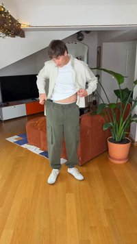 Žalios darbo kelnės su elastingais apačios kraštais ir šoniniais kišenėmis, derinamos su baltu marškinėliu ir dryžuota, šviesios spalvos palaidine.