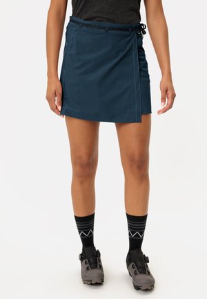 Marineblauwe wikkelrok met een gladde textuur, voorzien van een zijsluiting en contrasterende stiksels. Gecombineerd met zwarte sokken en grijze schoenen.