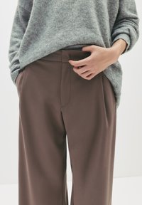 Personne portant un pantalon marron ample et un pull gris, ajustant la taille avec une main, se tenant contre un fond uni.