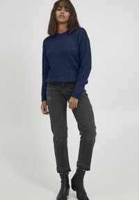 Marineblaues Strickpullover mit gerippten Manschetten, kombiniert mit schwarzen, hochtaillierten Jeans mit rohen Säumen und spitzen schwarzen Stiefeletten.