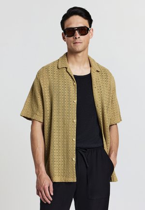 Camicia leggera khaki a maniche corte in pizzo con bottone frontale e colletto aperto, indossata sopra un top nero texturizzato e pantaloni neri.