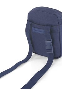 Bolso bandolera de tela azul marino con correas ajustables, cierre de cremallera y un parche de logo visible en la parte frontal. Forma compacta y rectangular.
