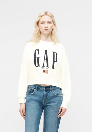 Ξανθιά γυναίκα που φοράει λευκό φούτερ GAP με λογότυπο και μπλε τζιν, στέκεται μπροστά σε απλό φωτεινό φόντο.
