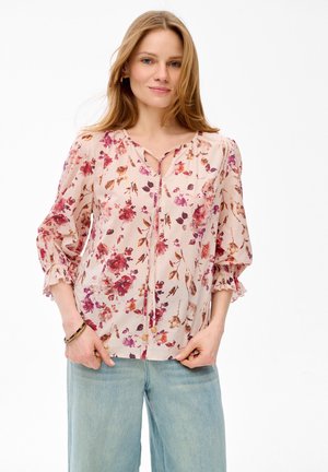 Woman wearing light pink floral blouse with 3/4 puffed sleeves and light blue jeans, standing with hands in pockets against white background.