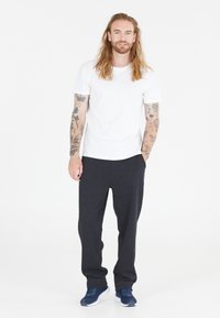 Vit kortärmad t-shirt, mörkgrå träningsbyxor, marinblå sneakers. Modellen har långt hår och tatueringar på armarna, stående mot en vit bakgrund.