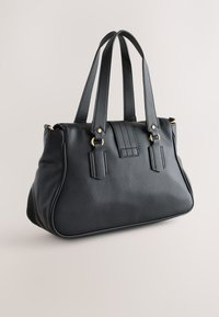 Sac à main en cuir noir texturé avec doubles poignées sur le dessus, anneaux métalliques dorés et forme rectangulaire structurée sur un fond neutre.