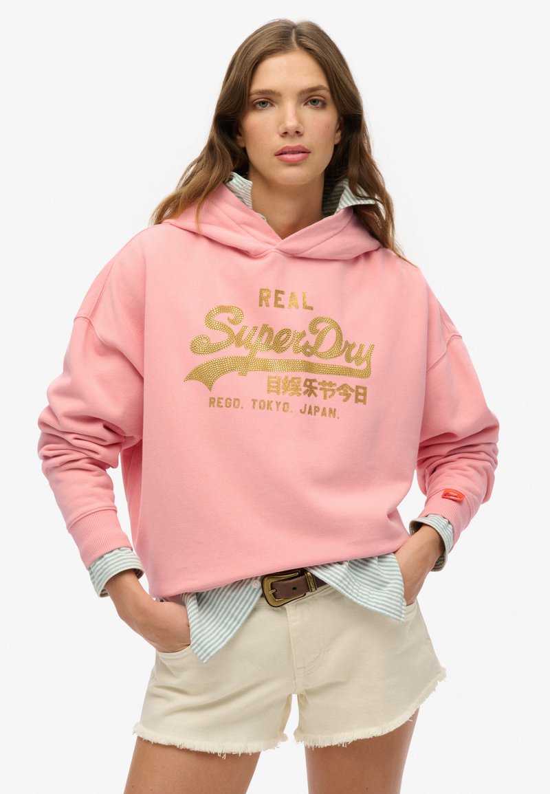 Felpa con cappuccio rosa con testo dorato "REAL SuperDry" e logo. Indossata sopra una camicia a righe blu e bianca, abbinata a pantaloni corti chiari.