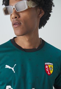 Grön kortärmad sporttröja med svart krage, Puma-logotyp och Racing Club de Lens-patch. Har subtila mönster och en välventilerad textur.