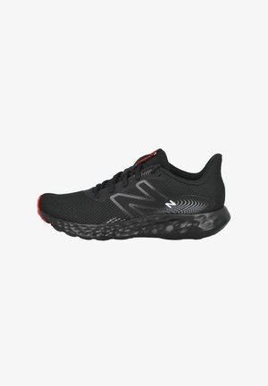 Zapatilla deportiva negra con parte superior de malla transpirable, cuello acolchado y suela negra texturizada con acentos rojos. Diseño de logo notable en gris.
