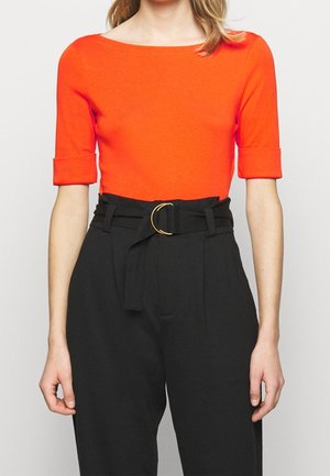 Vrouw draagt een feloranje top met ellebooglange mouwen, ingestopt in een zwarte broek met hoge taille, voorzien van een stoffen riem en een gouden gesp.