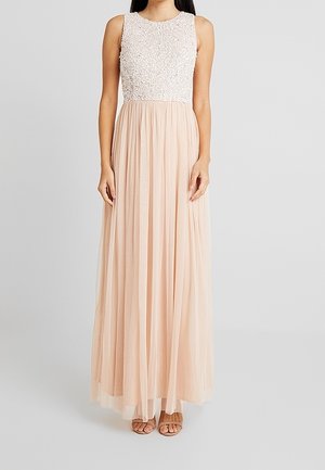 Suknia bez rękawów do ziemi z fakturowanym białym gorsetem i zwiewną, jasno brzoskwiniową plisowaną spódnicą, noszona z sandałami w kolorze nude.