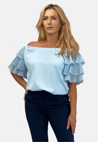 Jasnoniebieska bluzka bez ramiączek z warstwowymi falbankami, gładkiego materiału i luźnym kroju. Połączona z ciemnoniebieskimi spodniami.