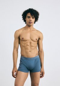 Boxeri scurți albaștri dintr-un material moale, cu o potrivire strânsă și un brâu gri Calvin Klein cu detalii ale logo-ului. Modelul stă cu brațele relaxate.