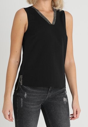 Mouwloze zwarte top met zilveren bies op de V-hals en een zijsplit, gedragen met vervaagde zwarte versleten jeans.