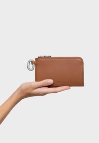 Cartera marrón de cuero con textura suave, cierre de cremallera y un adorno de mosquetón plateado; sostenida en una mano contra un fondo liso.