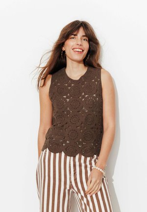 Femme souriante aux longs cheveux bruns porte un top au crochet marron sans manches et un pantalon blanc taille haute à rayures verticales marron devant un mur blanc.