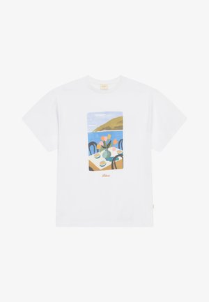 White cotton t-shirt featuring a graphic print of a table with flowers and cups, set against a coastal landscape background.