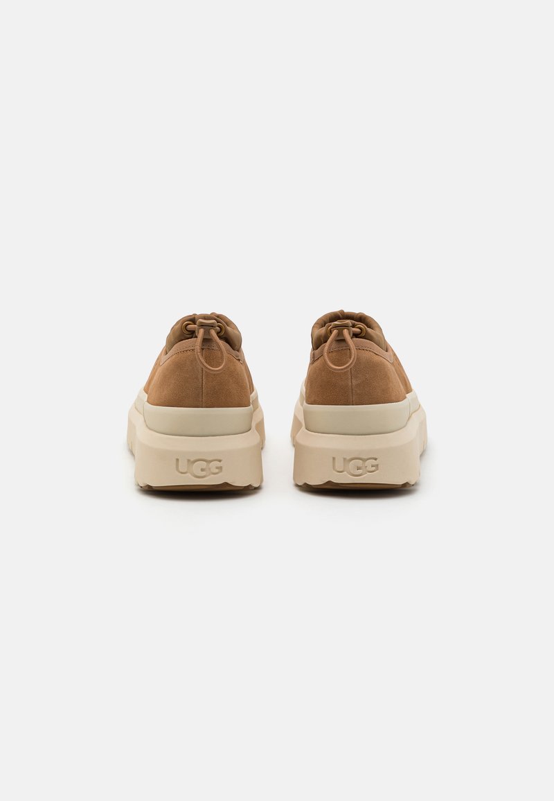Braune Wildlederschuhe mit einer beigen Gummisohle, runder Form und elastischen Schnürsenkeln, verziert mit dem UGG-Logo auf der Rückseite.