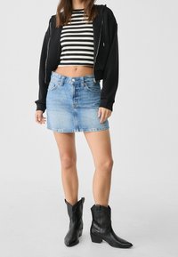 Sudadera negra con cremallera sobre un top corto a rayas blancas y negras, combinada con una minifalda de mezclilla azul claro y botas vaqueras negras.