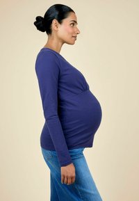 Top de manga larga morado con cintura fruncida, combinado con vaqueros de un azul claro. Mujer de pie de lado, mostrando una prominente barriga de embarazo.