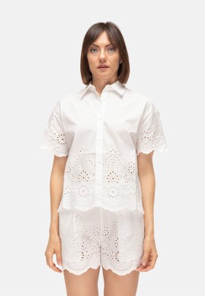 Donna che indossa una camicia bianca a maniche corte con bottoni e pantaloncini abbinati con ricamo floreale a occhielli e bordi smerlati.