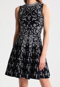 Svart ärmlös klänning med vita blommönster, figurnära liv, och vidkjol. Rund halsringning och textil med struktur.