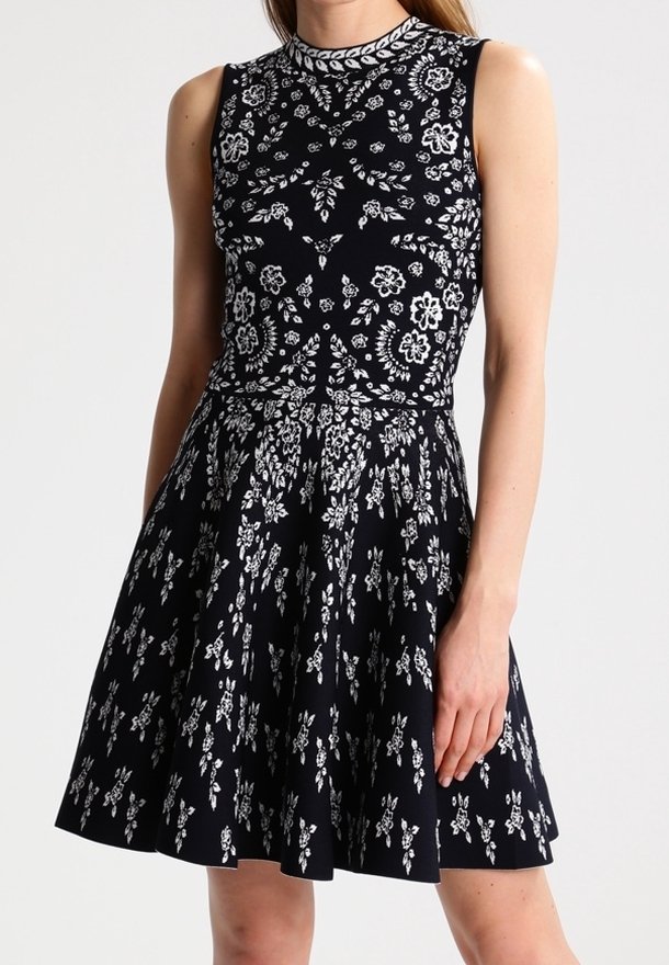 Svart ärmlös klänning med vita blommönster, figurnära liv, och vidkjol. Rund halsringning och textil med struktur.