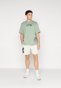 Φωτεινό πράσινο oversized μπλουζάκι με γραφικά σχέδια, σε συνδυασμό με κρεμ σορτσάκι που έχει μαύρες λεπτομέρειες. Το σύνολο ολοκληρώνεται με λευκά αθλητικά παπούτσια.