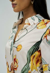 Blusa floreale con sfondo crema, motivi di fiori gialli e arancioni, colletto e bottoni, realizzata in tessuto morbido.