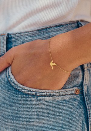Goldener Armreif mit einem minimalistischen Design in Form eines High Heels, mit einer zarten Kette und einer glatten Textur, getragen am Handgelenk nahe einer Jeans-Tasche.