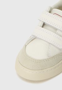 Hvit lær sneakers med beige suede-detaljer og borrelåsremmer. Har en teksturert gummisåle og sydd detaljer rundt kanten.