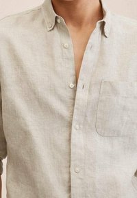 Chemise en lin beige clair avec col à boutons, poche avant et boutons visibles sur la patte de boutonnage ; tissu texturé et respirant.