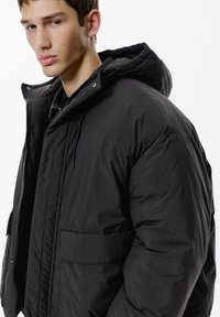 Jachetă puf din nylon neagră cu glugă, cu fermoar în față, buzunare mari cu clapetă și textură matlasată. Materialul pare ușor și durabil.