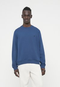 Μπλε βαμβακερό φούτερ με ριμπ μανσέτες και τελείωμα, στρογγυλή λαιμόκοψη, με μικρό λογότυπο στο στήθος· φοριέται με ανοιχτόχρωμο παντελόνι.