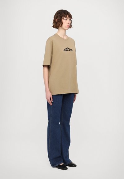Khaki oversized t-shirt met korte mouwen, ronde hals en zwart logo. Gecombineerd met blauwe wijde jeans en zwarte instapschoenen.