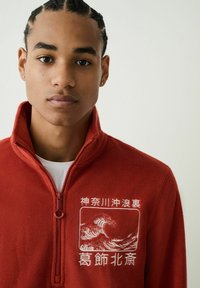 Piros polár cipzáras dzseki magas gallérral, fehér hullám mintás hímzéssel és sima textúrával. Cipzár részlet a nyaknál.