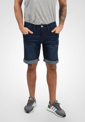 Shorts en denim bleu foncé avec un ourlet retroussé, design à cinq poches et fermeture à bouton. Portés avec un t-shirt gris et des baskets grises.