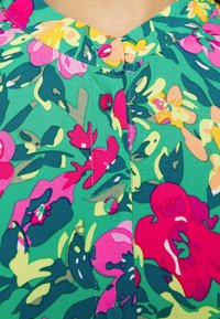 Blumenbluse mit grünem Hintergrund und leuchtenden rosa, gelben und blauen Blumen. Knopfdetails entlang des Ausschnitts. Weicher Stoff.