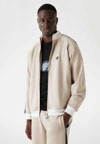 Beige trainingsjack met zwarte accenten, lichtgewicht stof, rits, geribde boorden, bijpassende broek, gedragen over een zwart graphic t-shirt.