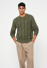 Olivenfarbener Kabelstrickpullover mit rundem Ausschnitt und gerippten Bündchen, kombiniert mit beigen Hosen. Kleines Logo-Patch am Saum.