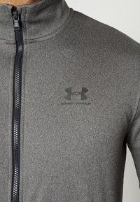 Chaqueta gris con cremallera y cuello alto, hecha de tejido texturizado. Presenta un logo negro en el pecho y una cremallera de contraste.