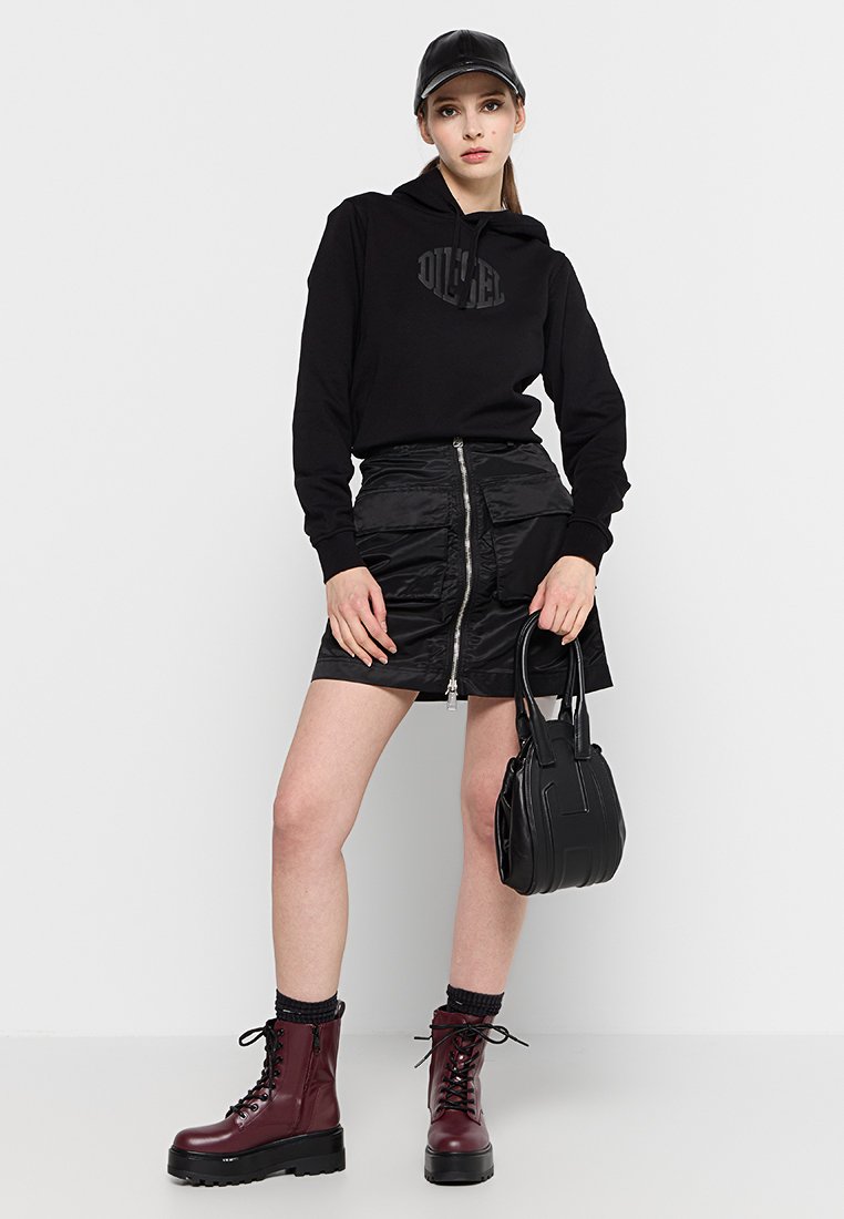 Haut à capuche noir avec logo, mini-jupe noire brillante avec fermeture éclair, bottes bordeaux à lacets et un petit sac à main noir.