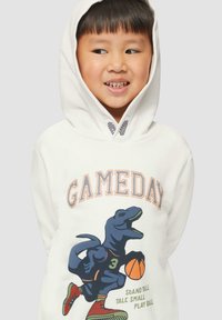 Biała bluza z kapturem z grafiką dinozaura w niebieskim kolorze trzymającego koszyk. Tekst "GAMEDAY" w pomarańczowym kolorze nad wzorem. Krój luźny z kapturem.