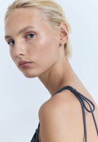 Portret al unei persoane cu păr blond deschis, îmbrăcată într-o ținută gri închis cu bretele subțiri, având o textură a pielii netedă și ochi albaștri.