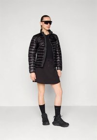 Chaqueta acolchada negra con acabado brillante, llevada sobre un top y falda negros. La modelo lleva gafas de sol negras, calcetines negros y zapatos negros gruesos.