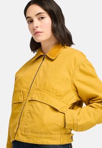 Yellow corduroy jacket featuring a zip closure, a collar, and two large front pockets. The fabric has a soft, textured appearance.