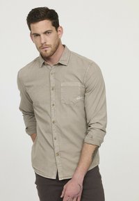 Chemise à manches longues beige clair avec un col boutonné, une poche sur le devant et des boutons marron. Texture lisse et coupe décontractée.