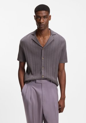 Hombre con una camisa de punto gris de manga corta y botones, con nervaduras verticales, y pantalones plisados de color púrpura claro, de pie con las manos parcialmente en los bolsillos.